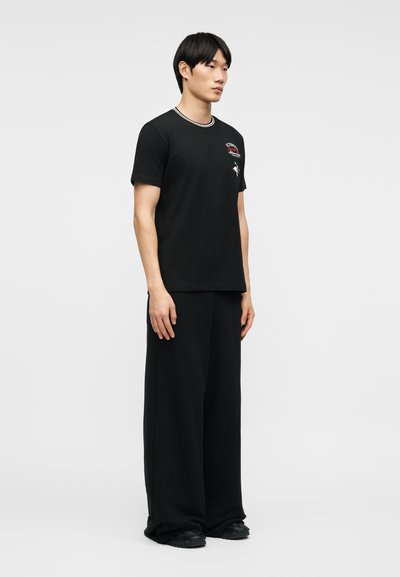 Νεαρός άνδρας που στέκεται, φοράει μαύρο κοντομάνικο μπλουζάκι με άσπρες ρίγες στον γιακά και φαρδιά μαύρα παντελόνια, μπροστά σε ένα απλό ανοιχτόχρωμο φόντο.