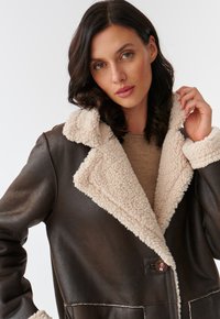 Veste en cuir marron avec un col et une doublure en fausse fourrure beige. Possède une fermeture à un bouton et des coutures blanches contrastantes.