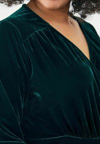 Grünes Samtkleid mit tiefem V-Ausschnitt, das über plissierte Details an den Schultern und eine glatte, weiche Textur verfügt.