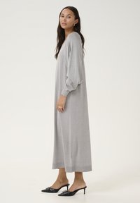 Robe longue grise avec une texture douce et scintillante, des manches bouffantes et une coupe ample, accompagnée de mules à talons noirs avec des accessoires décoratifs.
