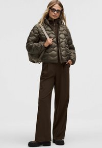 Kobieta ubrana w ciemnozieloną pikowaną kurtkę, dopasowane spodnie, czarne masywne buty, okulary przeciwsłoneczne oraz trzymająca beżową torbę na ramię na jednolitym tle.