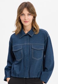 Denim jacket in dark blue with a collar, front zip, two chest pockets, and contrasting white stitching. Fitted design with a cropped hem.