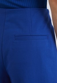 Pantalon bleu à la texture lisse, présentant une couture horizontale et une poche arrière unique avec un détail de rabat rectangulaire.