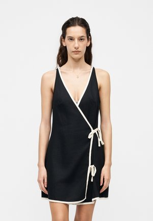 Hosszú sötét hajú nő ujjatlan fekete átkötős ruhát visel fehér szegéllyel és oldalsó megkötőkkel, sima világos háttér előtt áll.