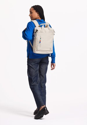 Beige backpack with a sleek, rectangular design, adjustable straps, a front pocket with zipper, and multi-colored pen holder accent.