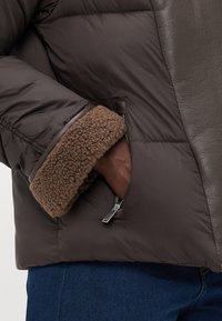Brun pufferjakke med lærdetaljer og fleecefôret mansjett. Har en glidelåslomme på siden for oppbevaring.