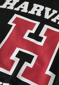 Juodas audinys su ryškiu raudonu ir baltu Harvardo "H" logotipu drąsiu šriftu, turinčiu siuvinėtą tekstūrą ir švarius kraštus.