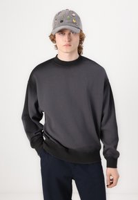 Crewneck sweatshirt in black, featuring long sleeves and a ribbed neckline. Paired with a gray cap adorned with colorful embroidered patches.