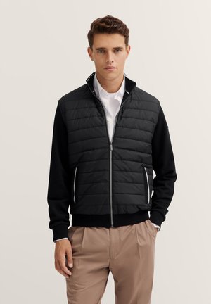 Junger Mann mit kurzen braunen Haaren trägt eine schwarze gesteppte Jacke mit Reißverschluss, ein weißes Hemd mit Kragen und hellbraune Hose und steht mit teilweise in den Taschen befindlichen Händen.