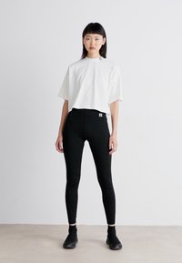 Balta trumpa marškinėliai su trumpomis rankovėmis, derinami su juodomis aukštu juosmeniu leggingais. Modelis vilki juodas kulkšnines batus su lygiu paviršiumi.