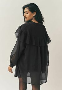 Fekete áttetsző ruha, hátul réteges fodros kialakítással, hosszú ujjakkal és bő szabással. Az anyag finom textúrájú.