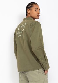 Olive green long-sleeve shirt with a collar, featuring a white graphic print on the back, and buttoned cuffs. Soft, smooth cotton fabric.