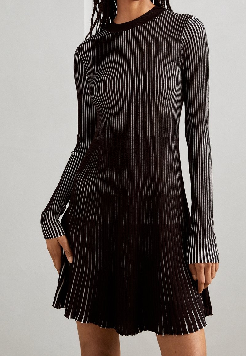 Femme portant une robe à manches longues rayée verticalement noire et blanche, avec un corsage ajusté et une jupe évasée.