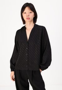 Black blouse with a V-neck and long, puffed sleeves. Features a textured design with diagonal stripes and polka dots. Button-front closure.