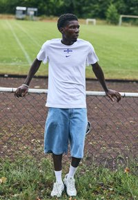 Weißes T-Shirt mit kurzen Ärmeln, blauer "ENG" Schrift und Nike-Logo, hellblaue Jeansshorts und weiße Sneakers. Steht an einem Zaun auf einem Rasenfeld.