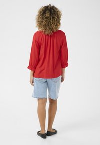 Rdeča bluzica z dolgimi rokavi in nabranimi detajli na hrbtu, v kombinaciji s svetlo modrimi jeans kratkimi hlačami in črnimi balerinkami. Preprost in neformalen dizajn.