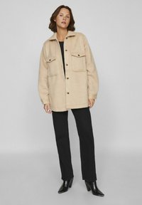 Vaaleanbeige villasekoite takki, jossa on kauluskäänne, napitus edessä, kaksi rintataskua ja pitkähihaiset hihat, yhdistettynä mustiin suoriin housuihin.