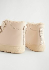Kurze beige Stiefel aus synthetischem Material mit einem flauschigen Innenfutter, ausgestattet mit einem glitzernden silbernen Akzent an der Rückseite und einer strukturierten Sohle.