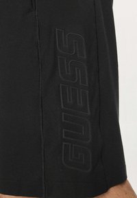 Pantaloncini neri in tessuto morbido, caratterizzati da un logo "GUESS" nero in rilievo ben visibile lungo il lato, con un design semplice e lineare.