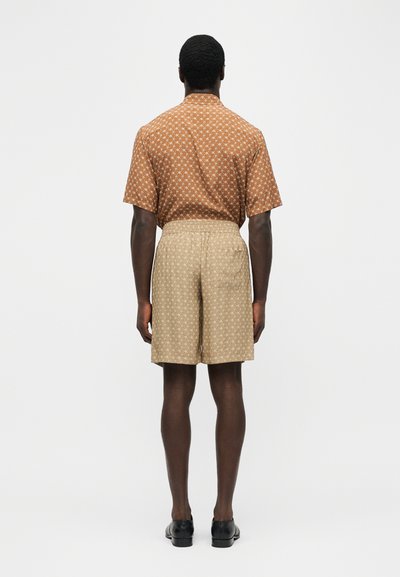 Homme debout, de dos, portant une chemise à manches courtes marron à motifs, un short beige à motifs et des chaussures habillées noires sur un fond uni.