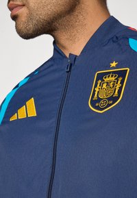 Chaqueta azul marino con cremallera que presenta un escudo de España dorado y un logo de Adidas amarillo. Incluye detalles en verde azulado y rojo en los hombros. Tejido texturizado.