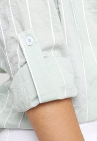 Helles grün gestreiftes Hemd mit weißen Akzenten. Verfügt über eine Knopfmanschette und eine zerknitterte Textur. Der Stoff wirkt leicht und atmungsaktiv.
