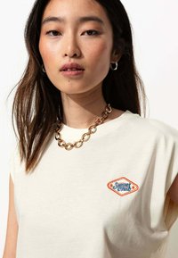 T-shirt en coton couleur crème avec des manches courtes et un patch brodé en forme de diamant "Sunset Club" en orange et bleu. Collier à maillons en or épais.