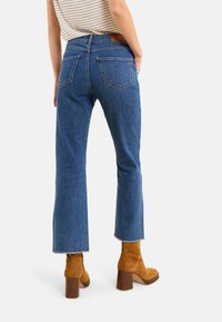 Jeans de denim azul con una cintura alta, corte acampanado, dobladillo desgastado y bolsillos traseros, combinados con botines de tacón bloque en color marrón claro.