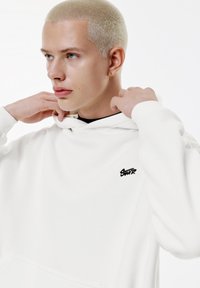 Vit hoodie gjord av mjukt tyg med en framficka. Har en liten svart logotyp på bröstet och är utformad med en dragsko på huvan.