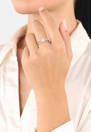 Mano con un anello di fidanzamento in argento che presenta un diamante centrale e pietre più piccole sulla fascia, tenuto vicino al volto contro una camicia bianca.