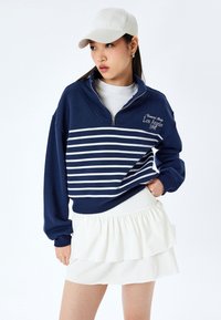 Sudadera azul marino con cremallera, rayas blancas y texto bordado, combinada con una falda blanca con volantes y una gorra de color claro.