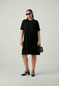 Μαύρο φόρεμα κοντό μανίκι με κλείσιμο από κουμπιά, υφασμάτινη υφή, φαρδιά σιλουέτα. Φοριέται με μαύρες γόβες και μια μικρή υφασμάτινη τσάντα.