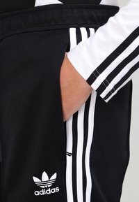 Černé sportovní kalhoty s bílým logem Adidas. Mají boční kapsu, černo-bílý proužek na vnějším švu a hladkou texturu.