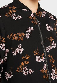 Svart jacka med rosa och bruna blommönster, med dragkedja fram och ribbad krage, bärs av en person från nacken och ner.