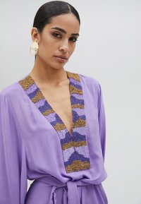 Ljubičasta bluzica s dubokim V-izrezom, ukrašena beaded dizajnom u zlatnim i plavim tonovima oko izreza, lagana tkanina, pojas u struku.