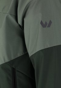 Groene jas met een gladde textuur en een tweekleurig ontwerp. Een logo in donkergrijs is zichtbaar op de schouder. Ritsdetails zijn zichtbaar aan de onderrand.