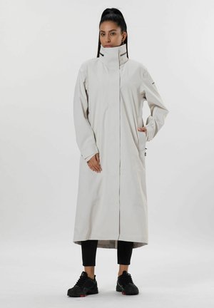 Lichtbeige waterdichte jas met hoge kraag, volledige rits aan de voorzijde en zijzakken. Wordt gedragen met zwarte leggings en schoeisel.