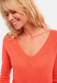 Femme souriante avec des cheveux blonds portant un pull en tricot à col V corail sur un fond clair.