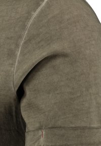 Camisa olive green de manga corta, hecha de una suave mezcla de algodón, con un detalle de costura y un pequeño acento de puntada roja cerca del dobladillo.