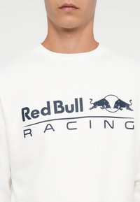 Férfi fehér pulóverben, amelyen a sötétkék "Red Bull Racing" logó látható, két egymással szembenéző bika fölött a szöveg felett.