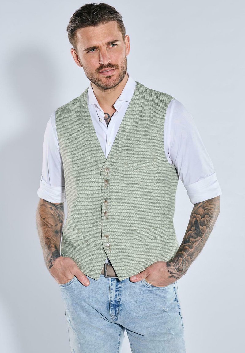 Mann mit Tätowierungen, der ein hellgrünes strukturiertes Weste über einem weißen hochgekrempelten Hemd und hellblauen Jeans trägt, steht mit den Händen in den Taschen.