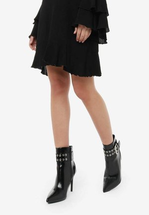 Bottines noires à bout pointu, avec un haut talon aiguille et des détails de sangle ornée de clous à la cheville. Matière en cuir brillant et texture lisse.