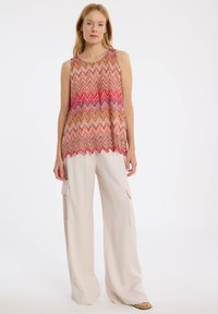 Mehrfarbiger ärmelloser Top mit Zickzackmuster in Rot, Rosa und Beige; der Saum hat einen wellenförmigen Schnitt. Kombiniert mit lockeren beigefarbenen Hosen.