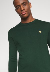 Maglione verde lavorato a maglia con scollo rotondo, caratterizzato da un logo di aquila ricamato in oro sul petto. Maniche lunghe e texture a coste.