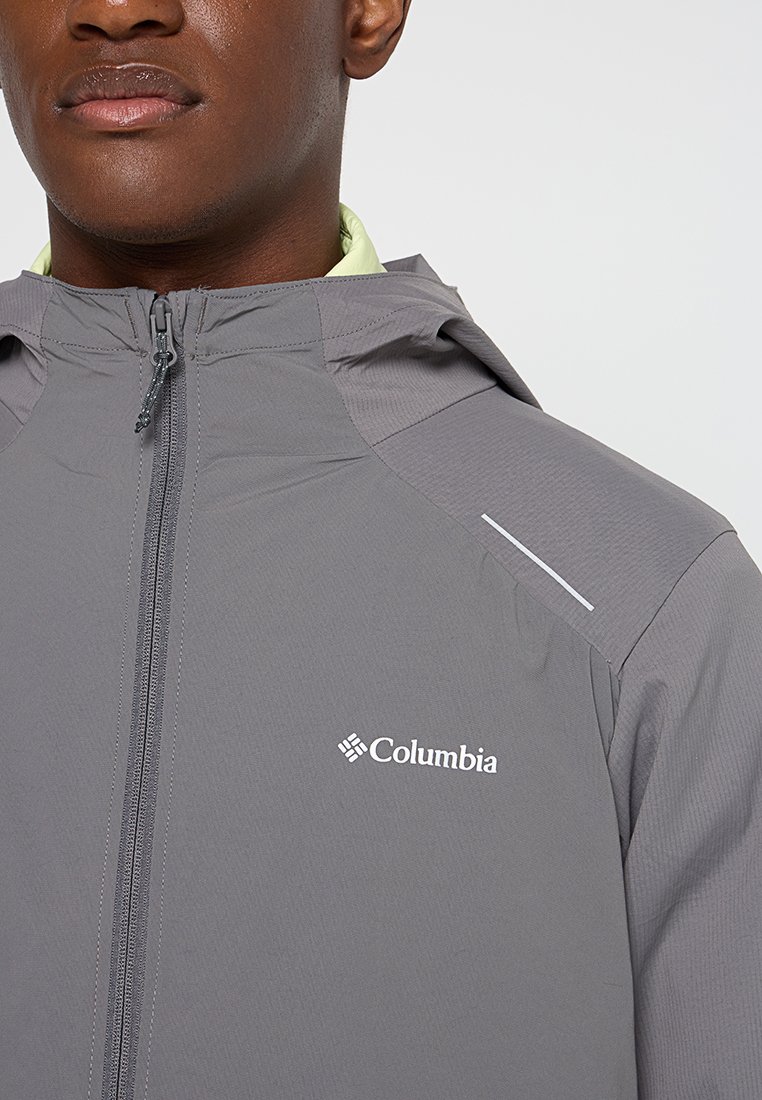 Nahaufnahme einer Person, die eine graue Columbia-Jacke mit einem Frontreißverschluss und Kapuze trägt, wobei das Markenlogo auf der Brust zu sehen ist.