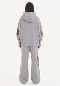 Grå hoodie med blommönster på ärmarna, i kombination med matchande sweatpants som har en färgglad grafik. Materialet verkar mjukt med en avslappnad passform.