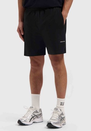 Person trägt schwarze Shorts mit elastischem Bund, weiße Socken mit Schriftzug und silberne Sportschuhe, steht vor einem schlichten Hintergrund.