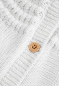 Tissu tricoté blanc avec des motifs torsadés et un bouton rond en bois gravé "NEXT" avec quatre trous.