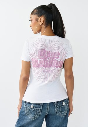 Kobieta z wysokim kucykiem, nosząca kolczyki koła, białą koszulkę z nadrukiem "True Religion" oraz niebieskie jeansy z detalicznymi tylnymi kieszeniami.