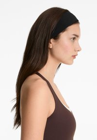 Svart elastisk huvudband som bärs av en modell, med slät yta och rund form, uppvisad mot en vit bakgrund. Modellen har långt brunt hår.